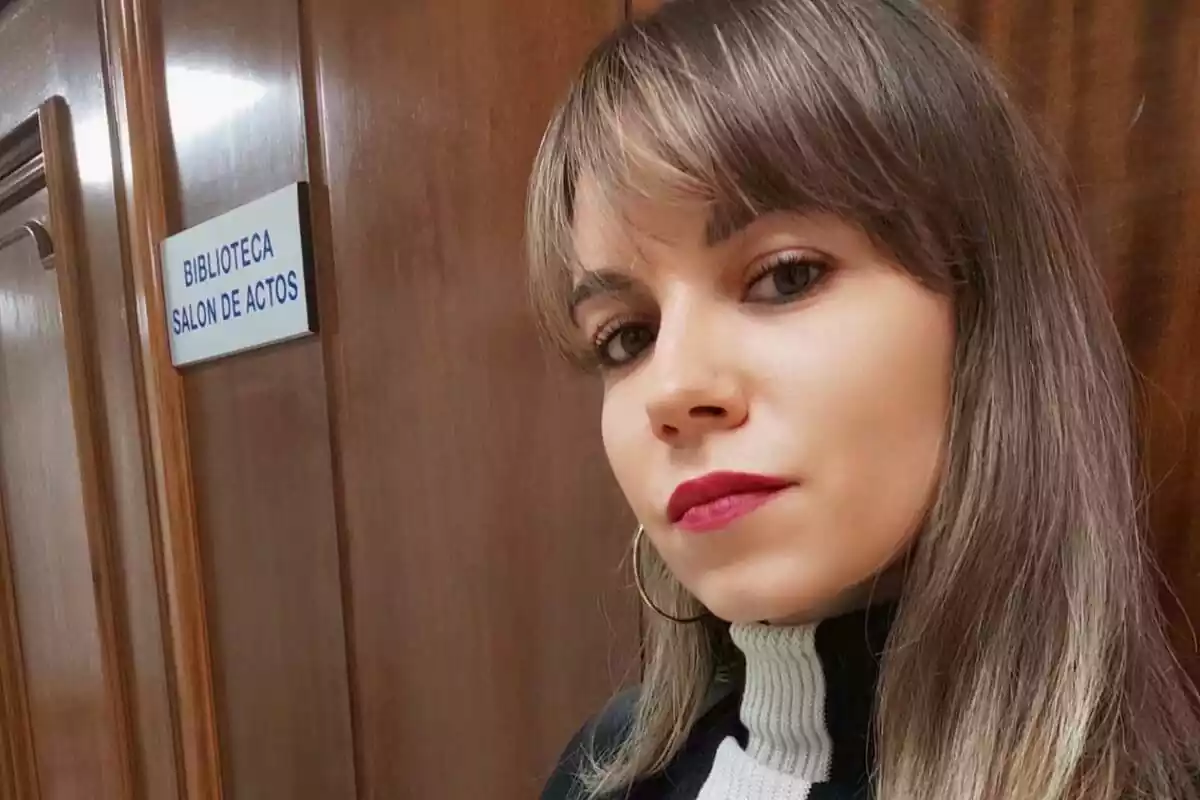 Primer plano de Paula Fraga haciéndose un selfie y de fondo una puerta de madera con un cartel que pone 'biblioteca salón de actos'