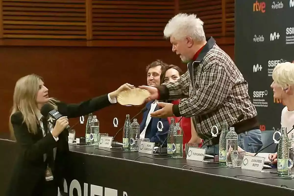 Paula Púa como reportera de Caiga quien Caiga entrega un objeto a Pedro Almodóvar durante una rueda de prensa, mientras otras personas observan desde la mesa.