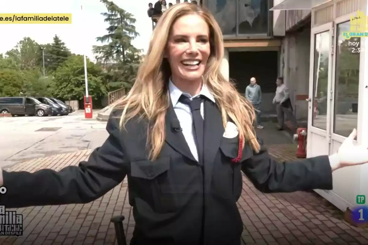Paula Vázquez sonriente con uniforme oscuro y corbata está al aire libre, con los brazos abiertos, mientras varias personas y vehículos se ven al fondo en el estreno de La Familia de la Tele. Paula Vázquez sonriente con uniforme oscuro y corbata está al aire libre, con los brazos abiertos, mientras varias personas y vehículos se ven al fondo en el estreno de La Familia de la Tele.