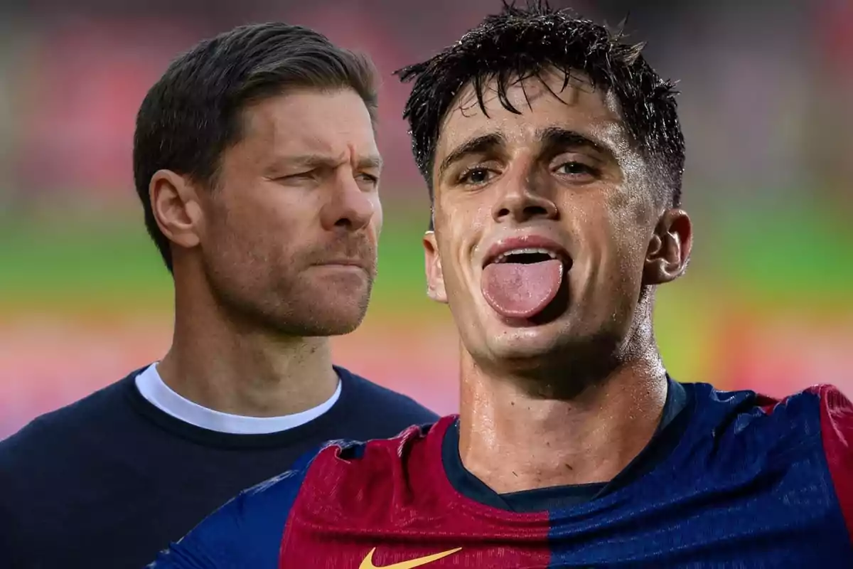 Un hombre con camiseta azul y roja saca la lengua mientras otro hombre de cabello corto y barba lo observa con expresión seria en un fondo desenfocado.