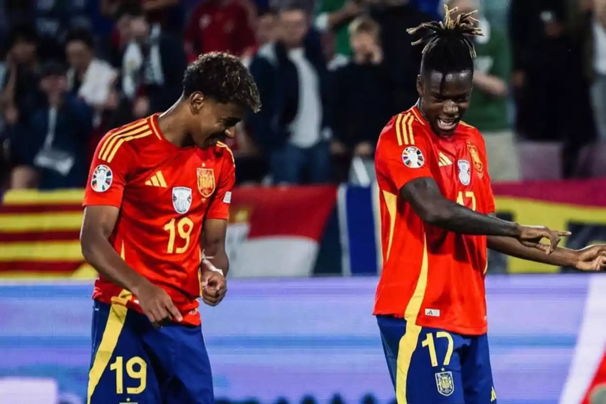 Dos jugadores de fútbol con uniformes rojos celebran en el campo mientras sonríen y señalan con las manos. Dos jugadores de fútbol con uniformes rojos celebran en el campo mientras sonríen y señalan con las manos.