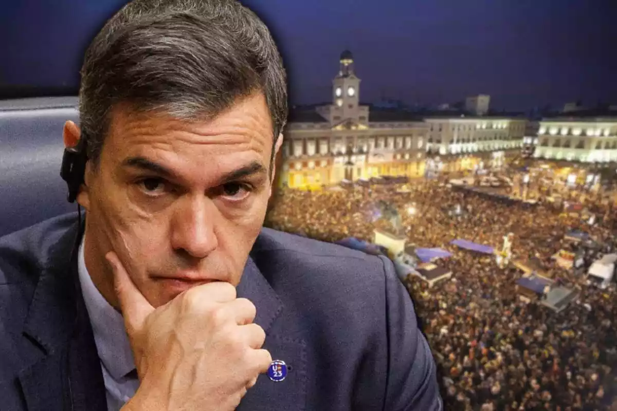 Plano medio corto de Pedro Sánchez con cara de preocupación y con su mano derecha en la barbilla y de fondo una imagen aerea de la acampada del 15M en la Puerta del Sol de Madrid de noche