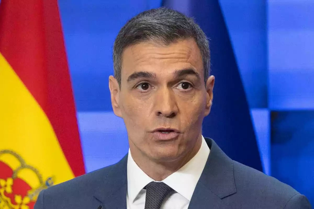 Hombre con traje y corbata hablando frente a una bandera de España y un fondo azul.