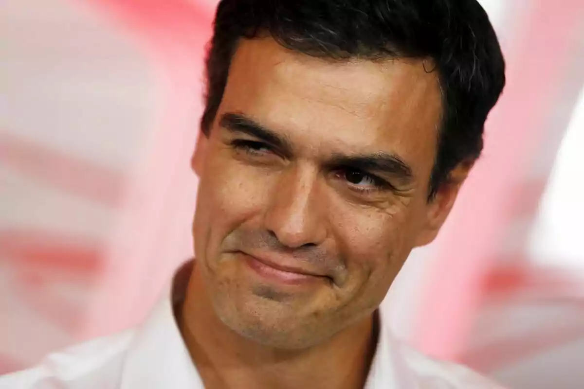 Hombre de cabello oscuro con camisa blanca sonriendo levemente frente a un fondo desenfocado en tonos rosados