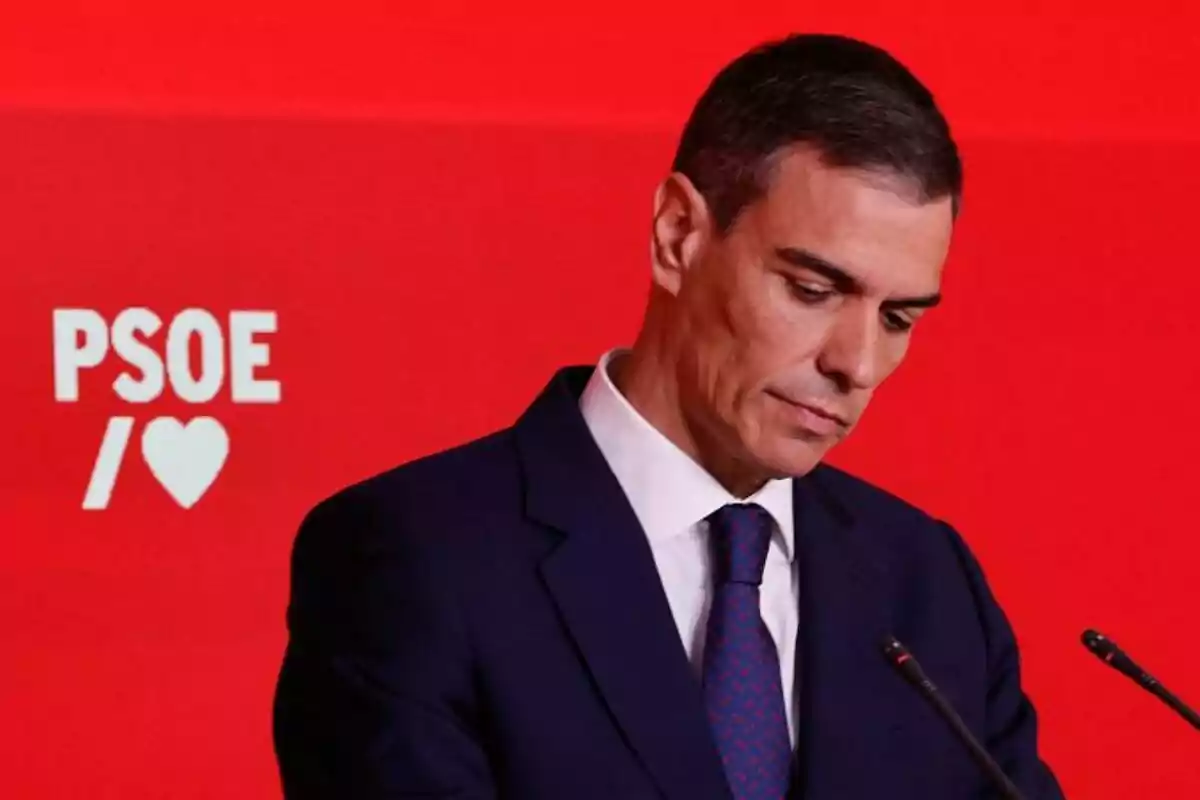 Hombre con traje oscuro y corbata azul frente a un fondo rojo con el logo de PSOE y un corazón blanco Hombre con traje oscuro y corbata azul frente a un fondo rojo con el logo de PSOE y un corazón blanco
