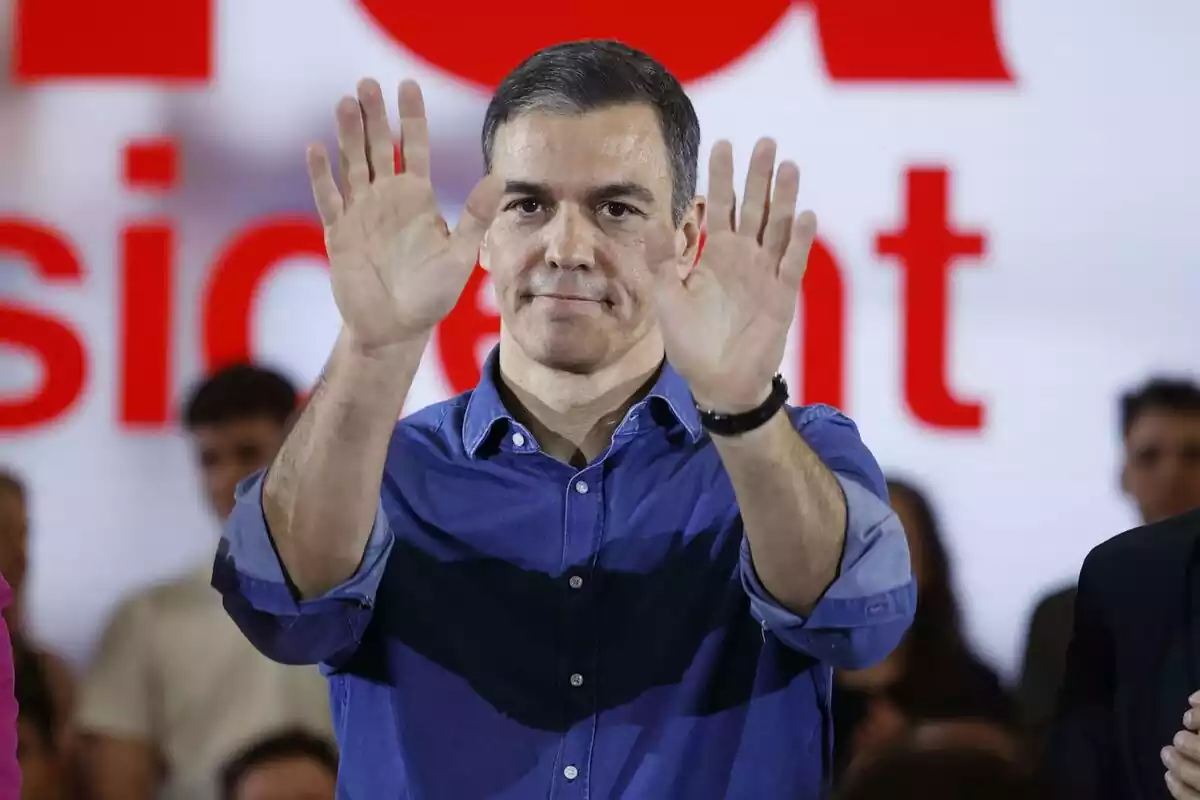 Plano medio de Pedro Sánchez en un mitin en Catalunya, con camisa azul y con los dos brazos y manos hacia adelante saludando