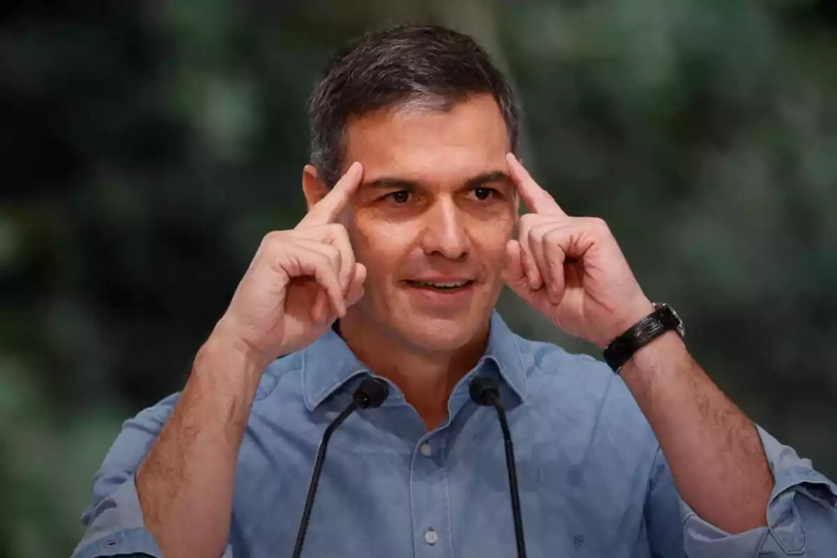 plano medio de Pedro Sánchez hablando delante de dos micrófonos con cara sonriente y con los dos dedos índice apoyados en su frente