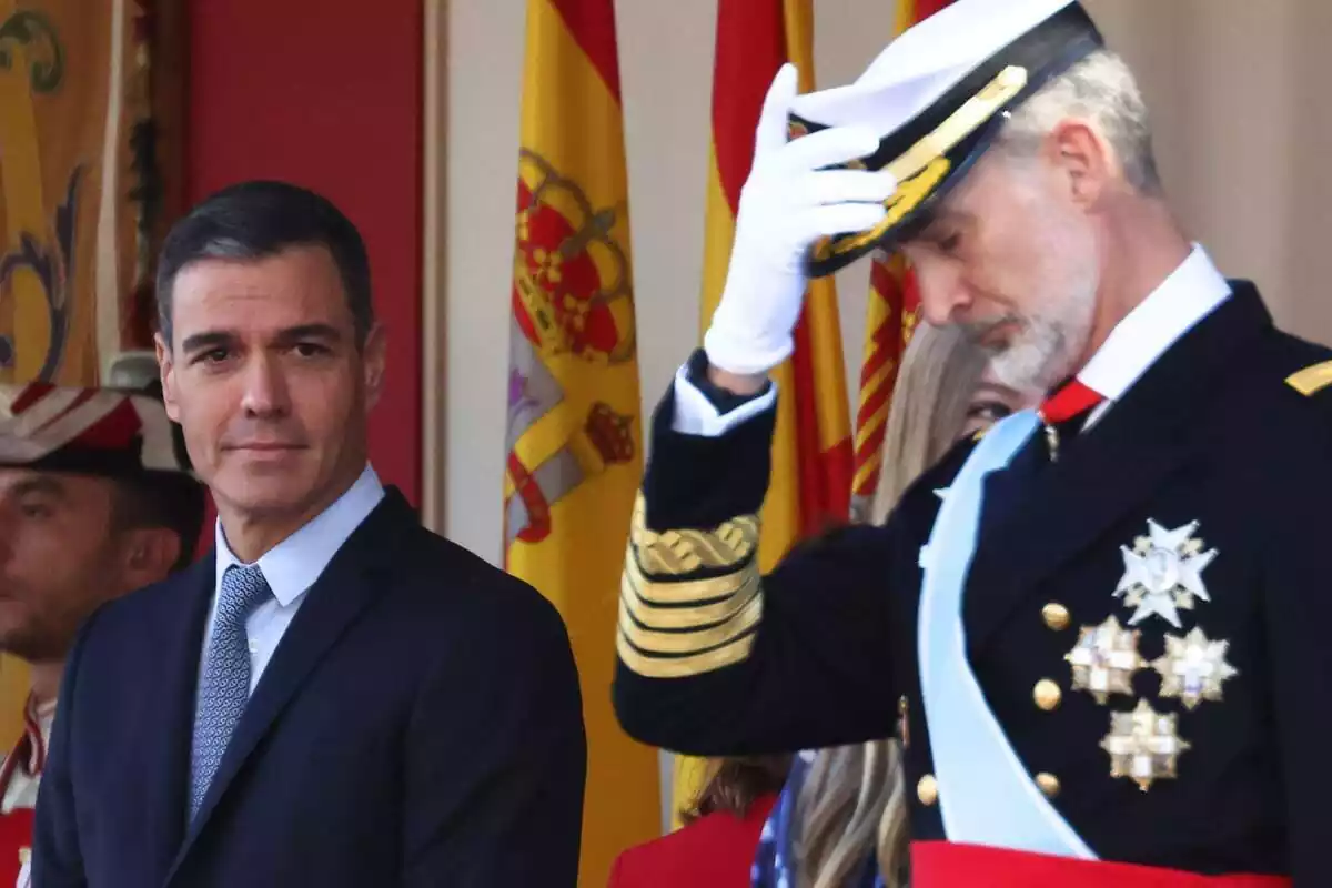 Plano medio de Pedro Sánchez con una ligera sonrisa mientras el rey Felipe vestido de militar se coloca bien la gorra mientras mira al suelo