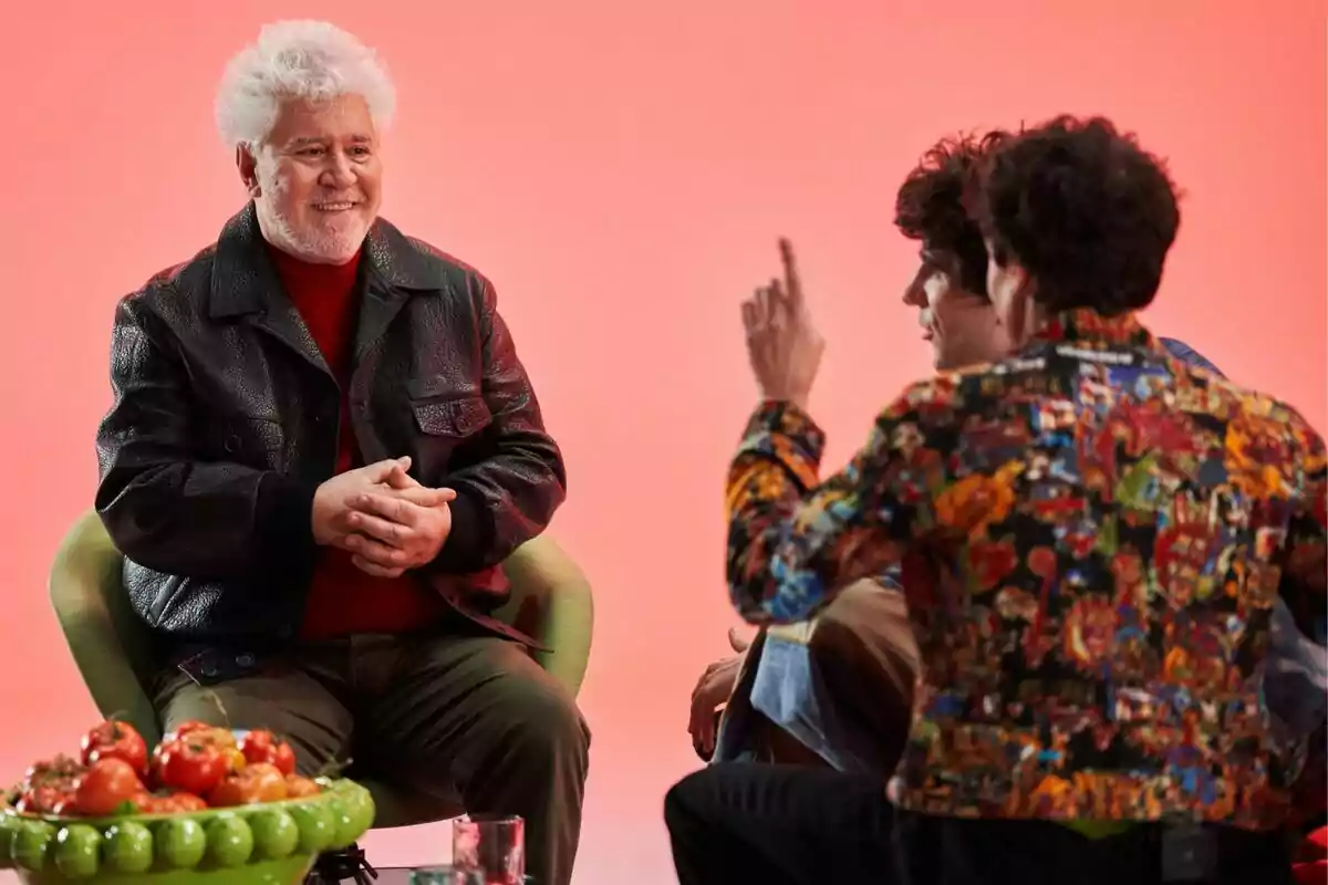 Pedro Almodóvar de cabello canoso y chaqueta negra sonríe mientras conversa con Javi Calvo y Javi Ambrossi frente a él, una de ellas lleva una chaqueta colorida y hay un frutero con tomates sobre la mesa, todo en un ambiente con fondo rosa en Pedro x Javis Pedro Almodóvar de cabello canoso y chaqueta negra sonríe mientras conversa con Javi Calvo y Javi Ambrossi frente a él, una de ellas lleva una chaqueta colorida y hay un frutero con tomates sobre la mesa, todo en un ambiente con fondo rosa en Pedro x Javis