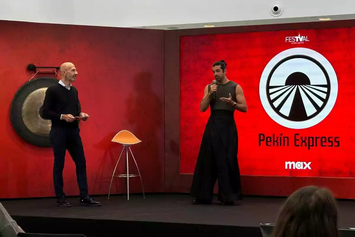 Miguel Ángel Muñoz y Alberto Carullo están de pie en un escenario con un fondo rojo, una de ellas sostiene un micrófono y la otra parece estar hablando, hay un logotipo que dice "Pekín Express" y un gong en la pared.