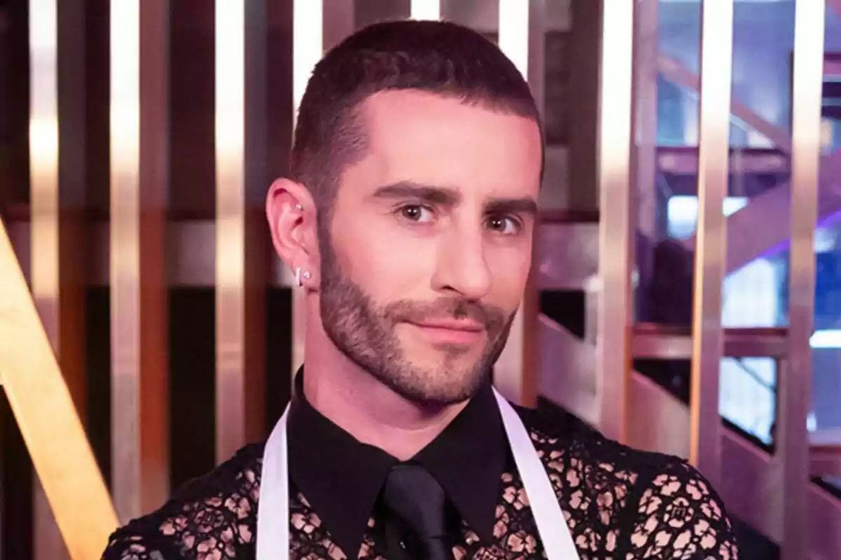 Pelayo Díaz con barba y cabello corto, usando una camisa de encaje negra y una corbata negra, con un delantal blanco, posando frente a una estructura metálica en MasterChef Celebrity.