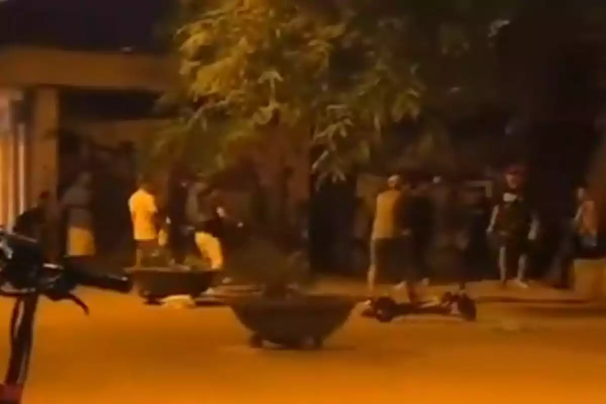 Un grupo de personas se encuentra reunido en una calle iluminada por luces amarillas, con macetas grandes y un árbol visible en el fondo.