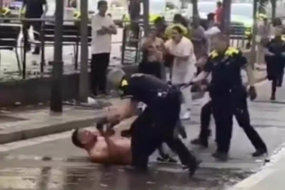 Un hombre sin camisa está siendo sometido en el suelo por un policía mientras otros oficiales y personas observan la escena en una calle.