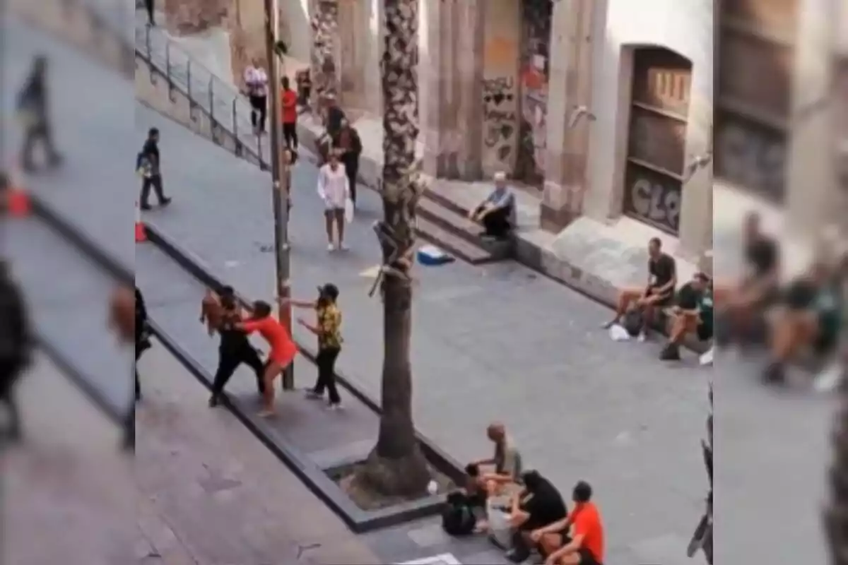 Una escena urbana muestra a varias personas interactuando en una calle, con algunos sentados en el suelo y otros de pie, mientras dos individuos parecen estar en una confrontación física cerca de una palmera.