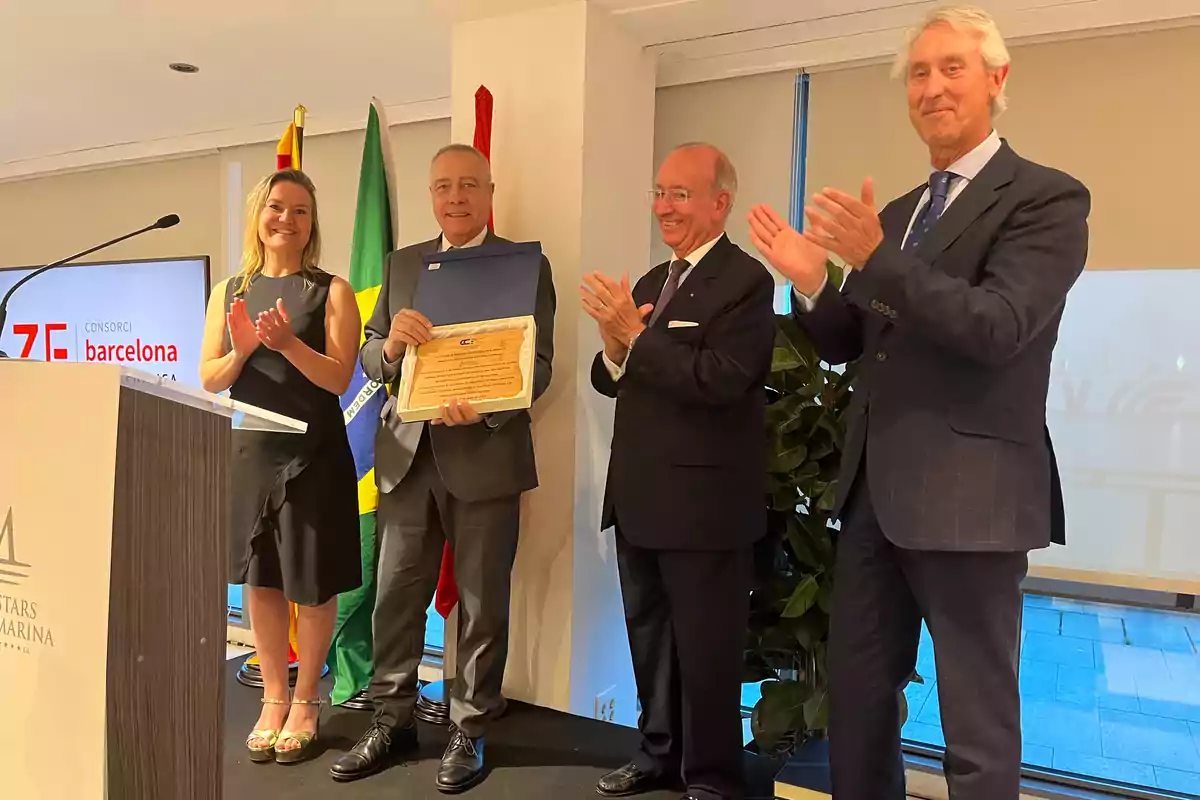 Un grupo de personas en un evento, una mujer y tres hombres, uno de ellos sosteniendo un diploma, todos aplaudiendo y sonriendo, con banderas y un podio en el fondo.