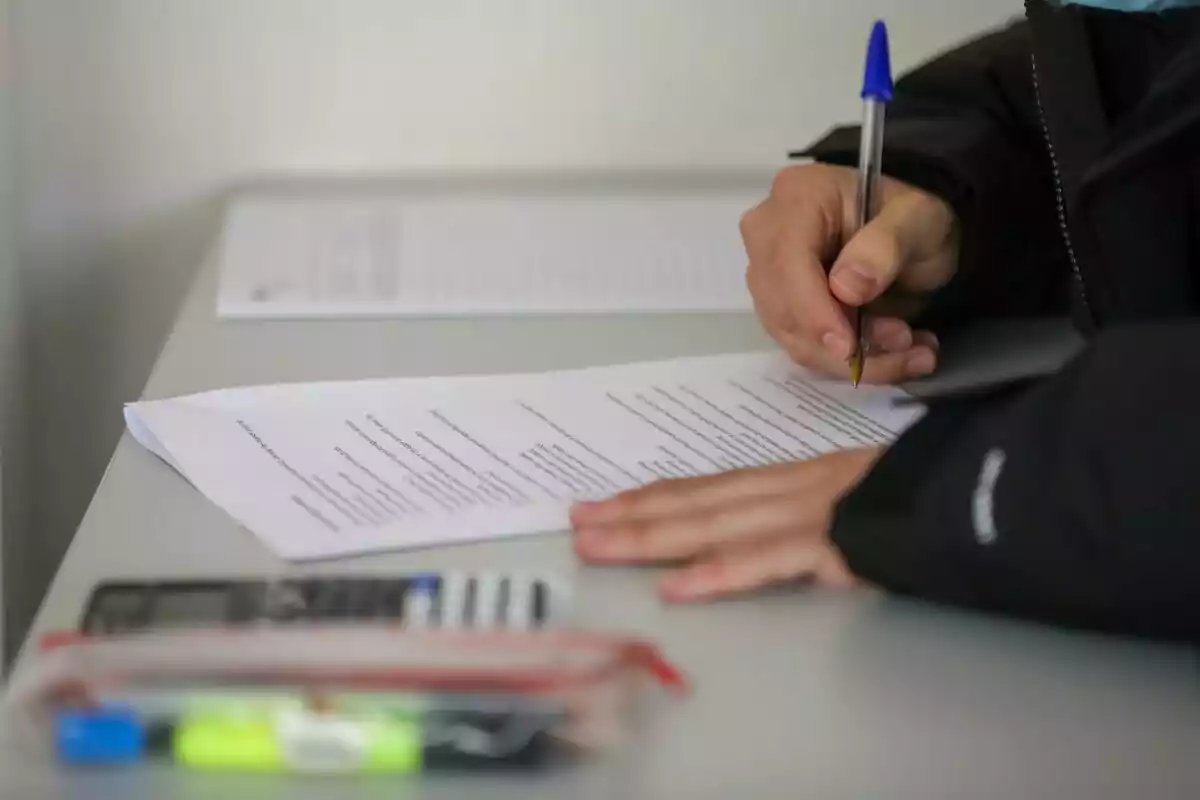 Una persona escribiendo en un documento sobre una mesa con un bolígrafo azul, junto a una calculadora y un estuche. Una persona escribiendo en un documento sobre una mesa con un bolígrafo azul, junto a una calculadora y un estuche.