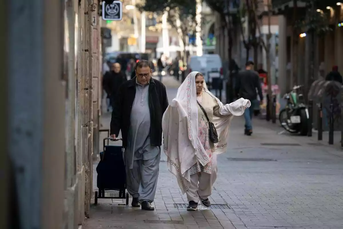 Una pareja de personas mayores caminando por una calle urbana, el hombre lleva una maleta con ruedas y la mujer viste un atuendo tradicional.