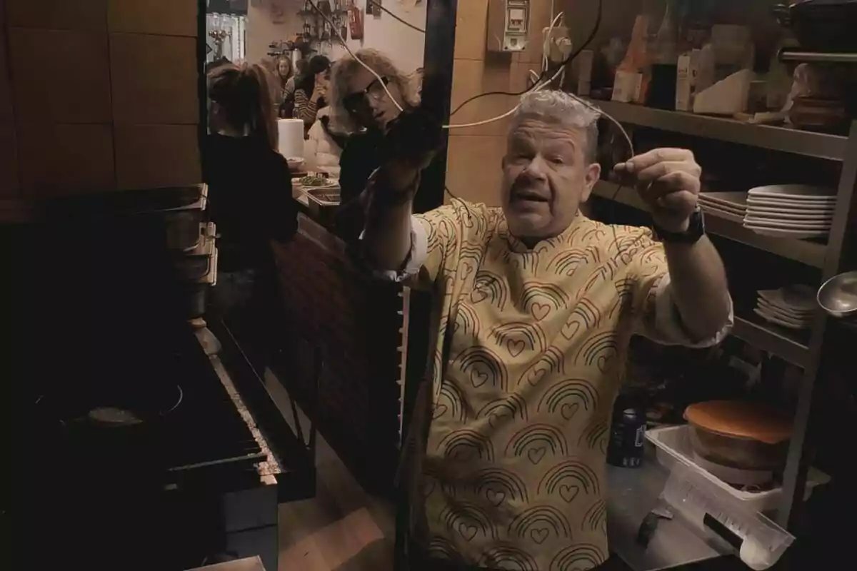 Alberto Chicote con una camisa colorida está en una cocina, sosteniendo utensilios de cocina, mientras varias personas están en el fondo, algunas sentadas y otras de pie, en lo que parece ser un restaurante o una cocina abierta en Pesadilla en la Cocina.