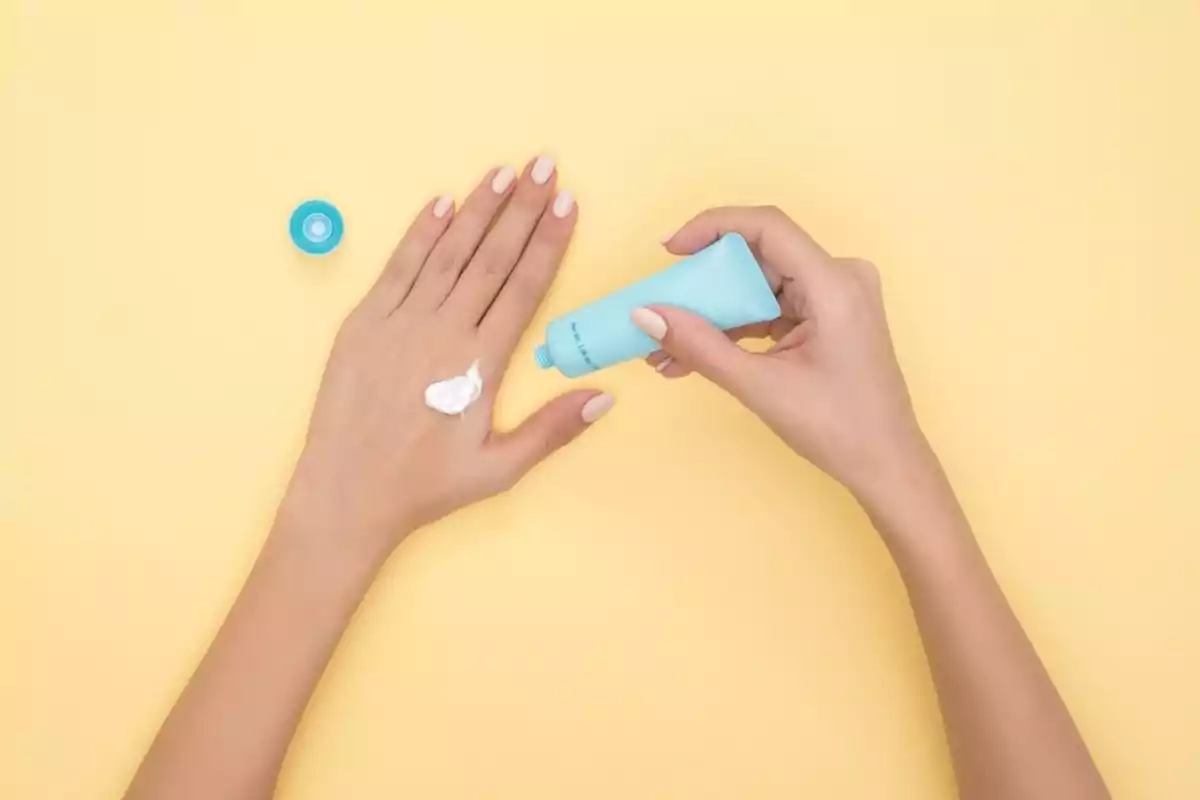 Manos aplicando crema de un tubo azul sobre un fondo amarillo.