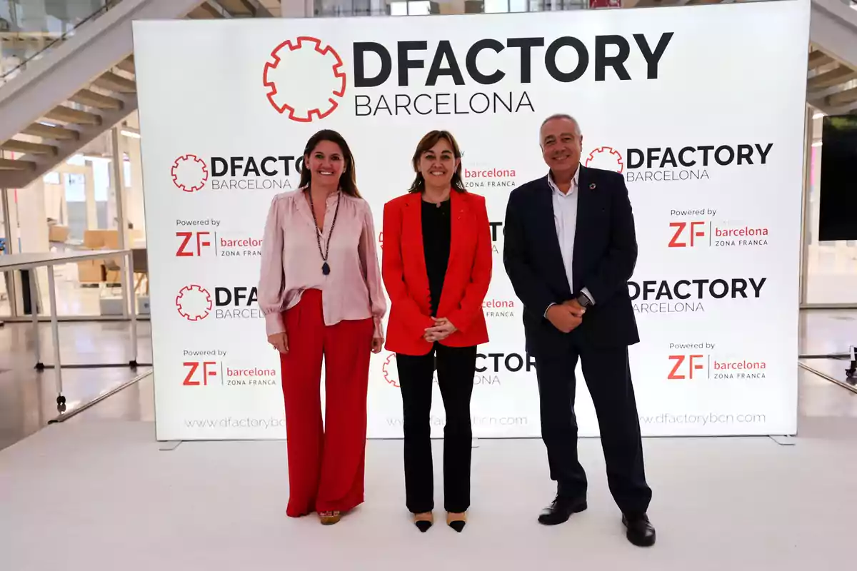 Tres personas posan frente a un cartel de DFactory Barcelona en un evento.