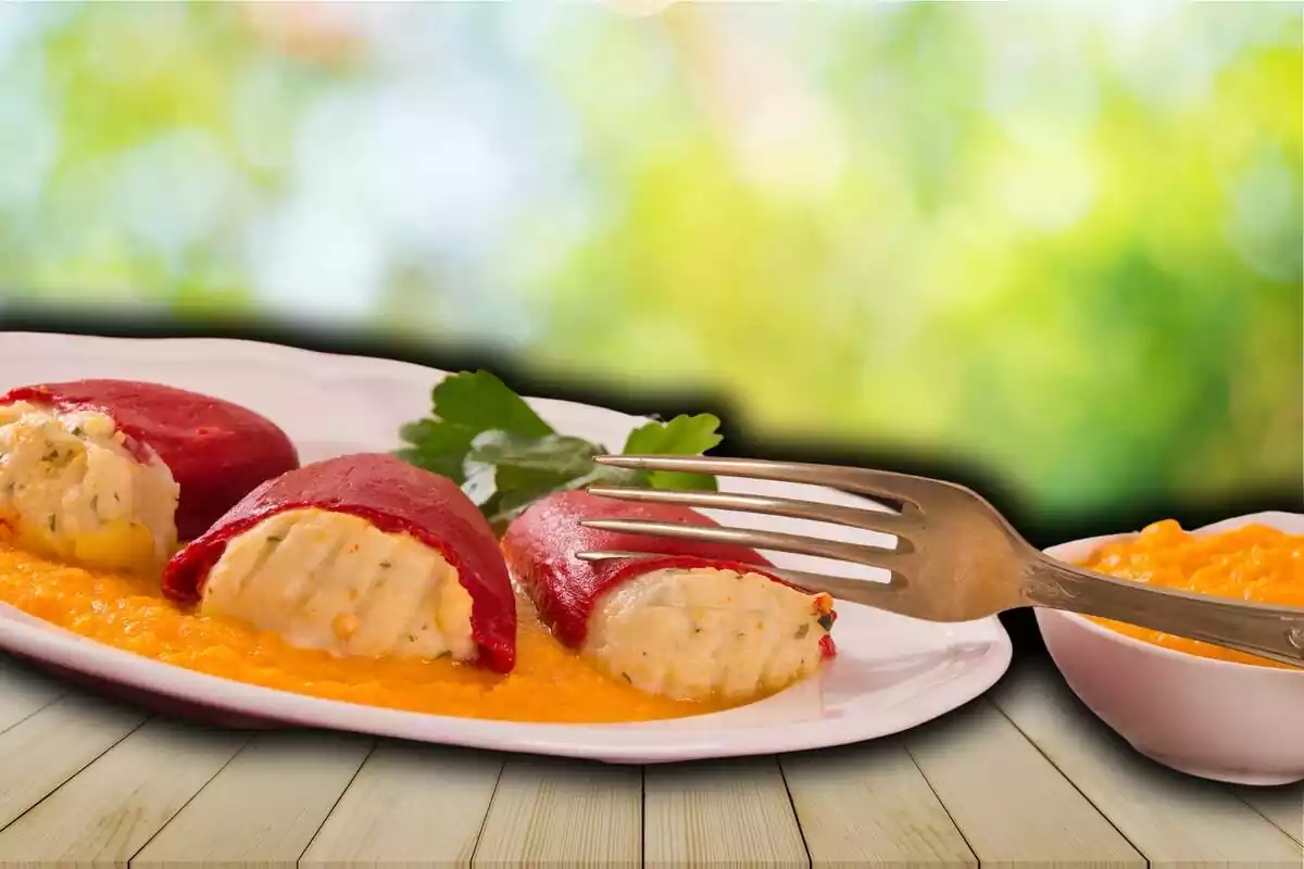 Montaje plato con pimientos rellenos de bacalao con un tenedor encima junto a un cuenco con más salsa sobre fondo de imagen de una mesa de madera en un jardín desenfocada