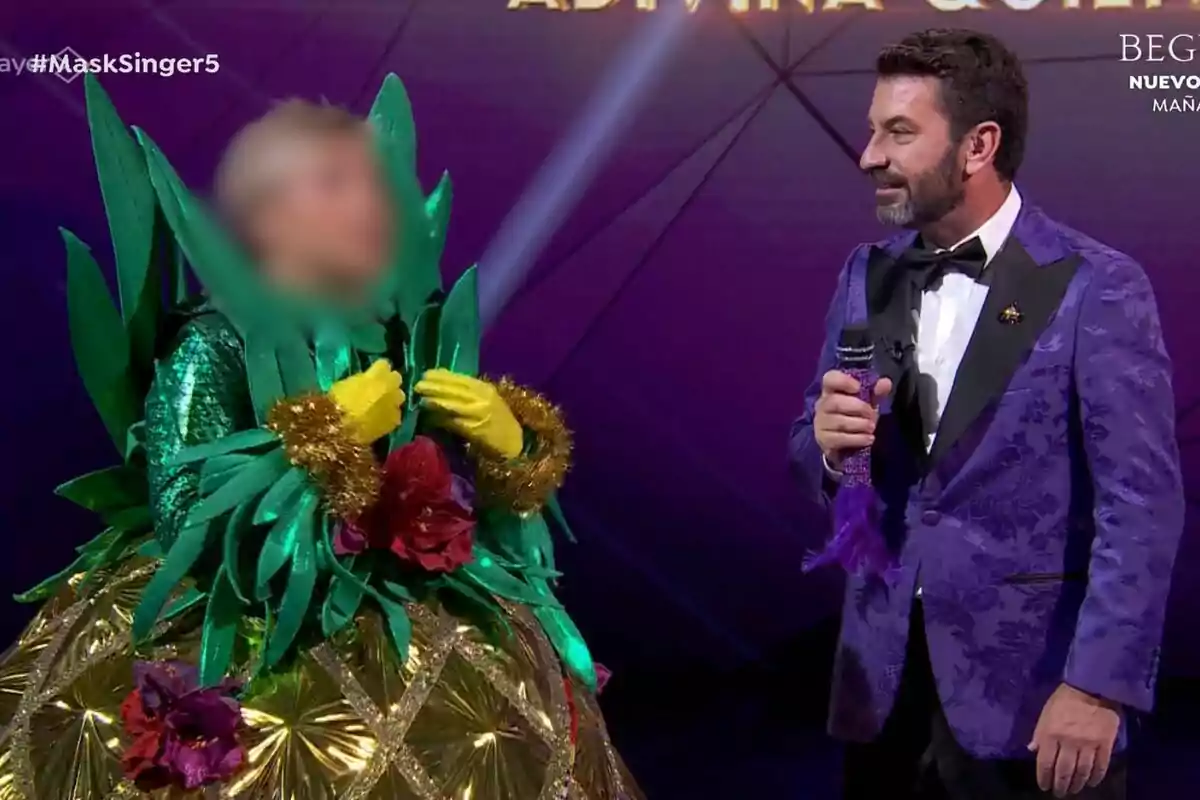 Una persona disfrazada de piña con hojas verdes y flores está junto a Arturo Valls que lleva un traje morado en un escenario de Mask Singer.