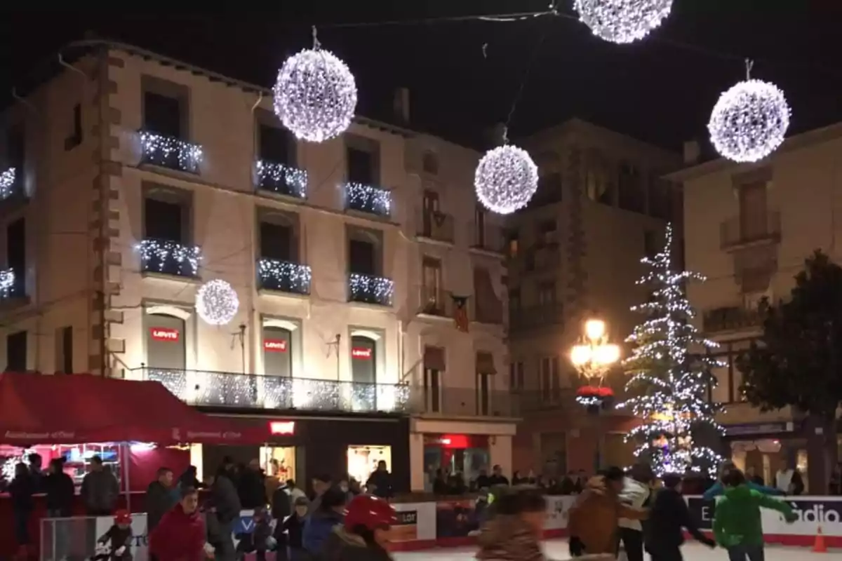 Personas patinando sobre hielo en una plaza decorada con luces navideñas y esferas brillantes colgantes.