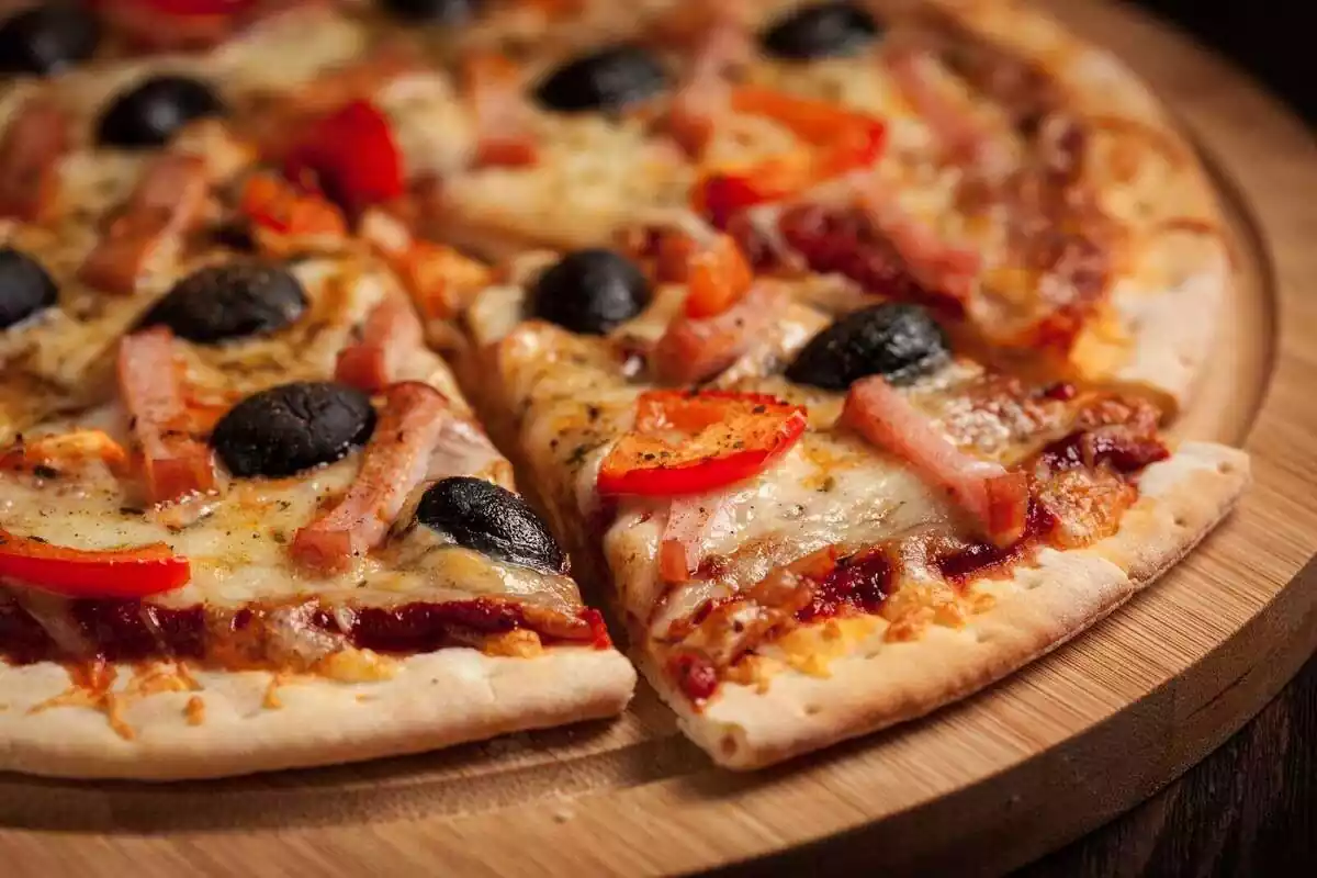 Primer plano de una pizza de jamón en trocitos con pimiento rojo y aceitunas en un tablero de madera sobre una mesa