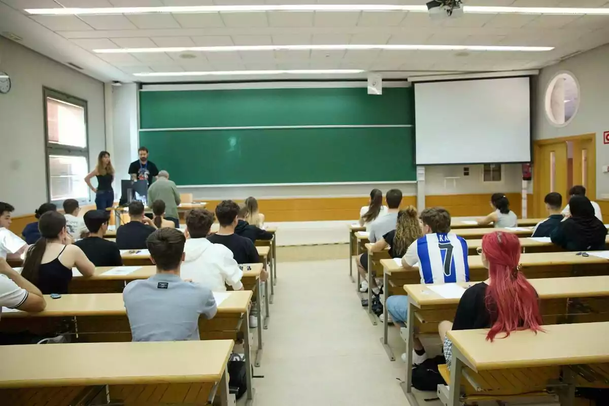 Aula universitaria con estudiantes sentados en pupitres mirando hacia el frente donde tres personas están de pie junto a una pizarra verde y una pantalla de proyección. Aula universitaria con estudiantes sentados en pupitres mirando hacia el frente donde tres personas están de pie junto a una pizarra verde y una pantalla de proyección.