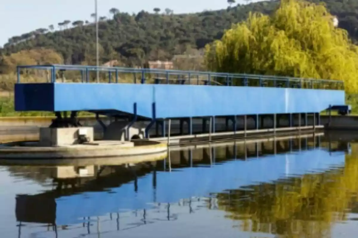 Una estructura azul sobre un cuerpo de agua con colinas y árboles al fondo.