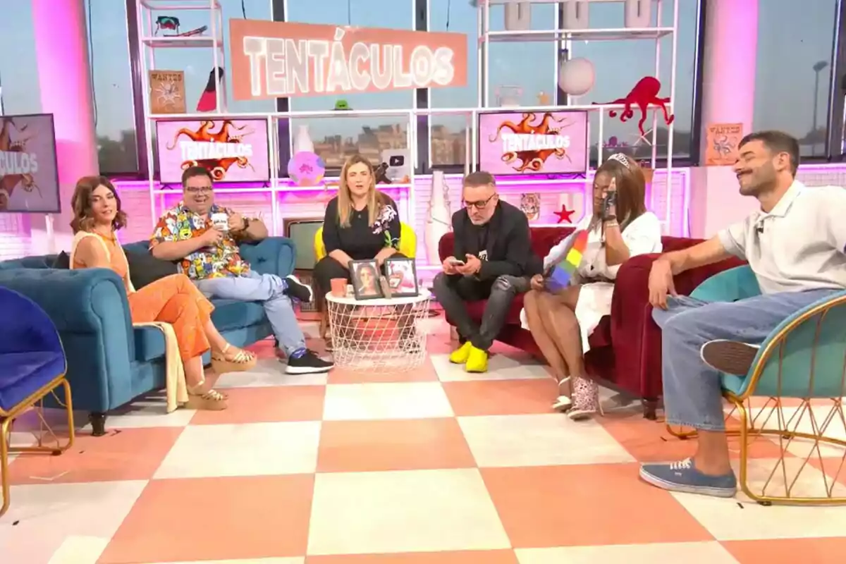 Seis personas están sentadas en un colorido set de televisión, 'Tentáculos', decorado con temática de tentáculos, conversando y sonriendo en un ambiente relajado. Seis personas están sentadas en un colorido set de televisión, 'Tentáculos', decorado con temática de tentáculos, conversando y sonriendo en un ambiente relajado.