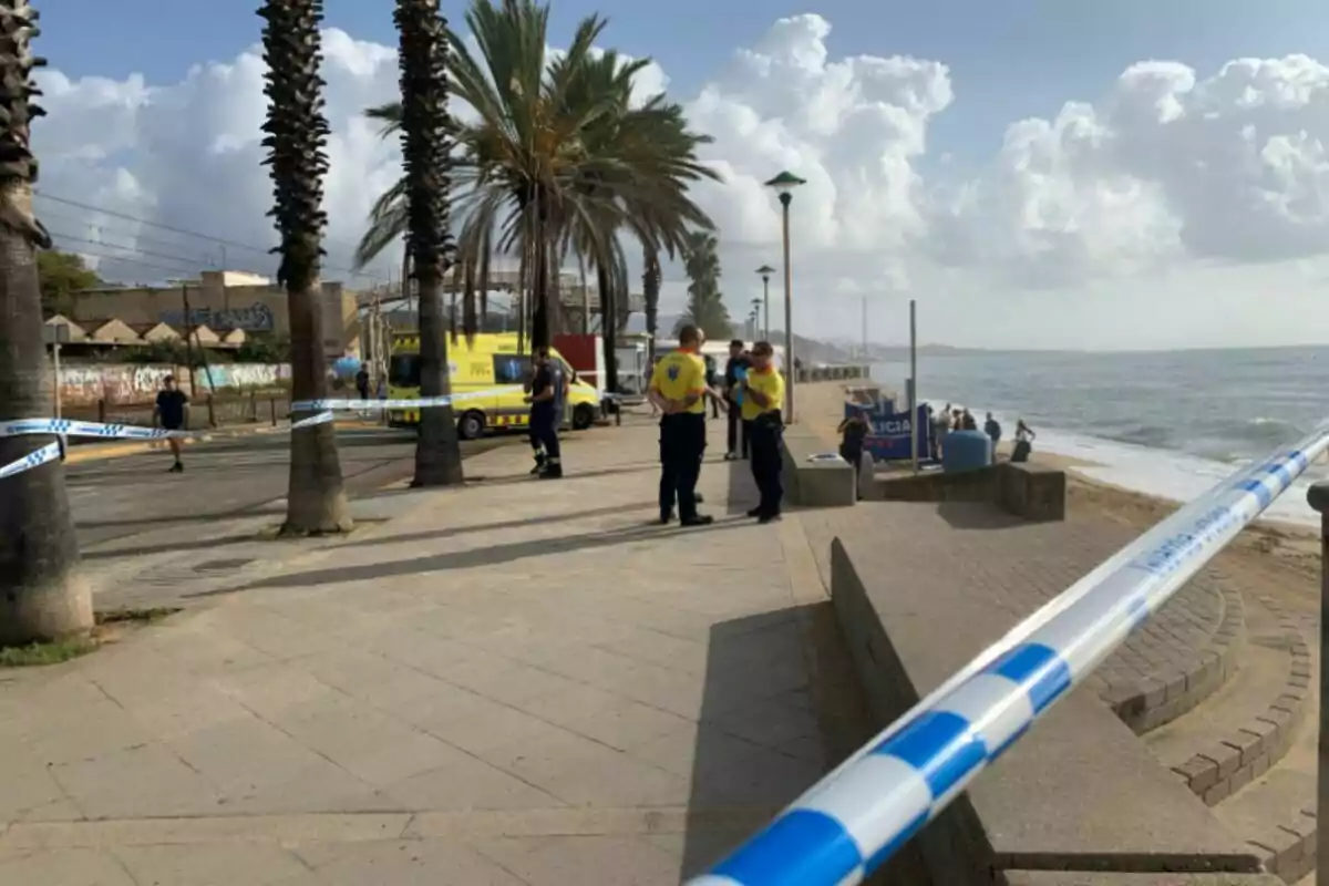 Una escena en una playa con varios policías y personal de emergencia, una ambulancia amarilla y cinta de seguridad delimitando el área. Una escena en una playa con varios policías y personal de emergencia, una ambulancia amarilla y cinta de seguridad delimitando el área.