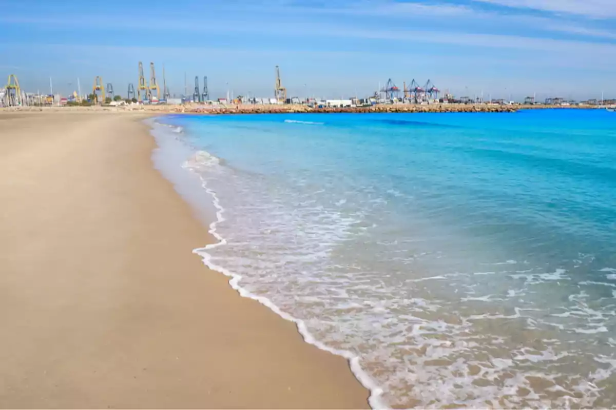 Playa de arena con agua azul y un puerto industrial al fondo bajo un cielo despejado. Playa de arena con agua azul y un puerto industrial al fondo bajo un cielo despejado.