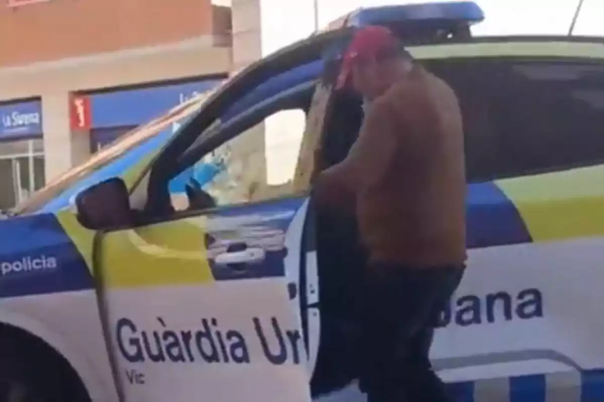 Un hombre con gorra roja está de pie junto a un coche de la Guàrdia Urbana de Vic, con la puerta del vehículo abierta.