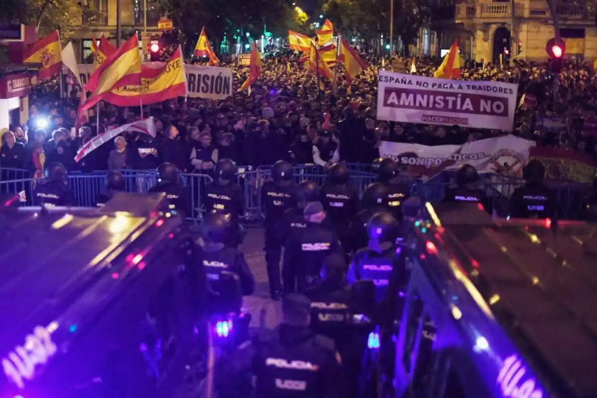 Plano general del cordón policial frente a la calle Ferraz de Madrid y delante, miles de manifestantes con banderas españolas y pancartas contra Pedro Sánchez y la amnistía