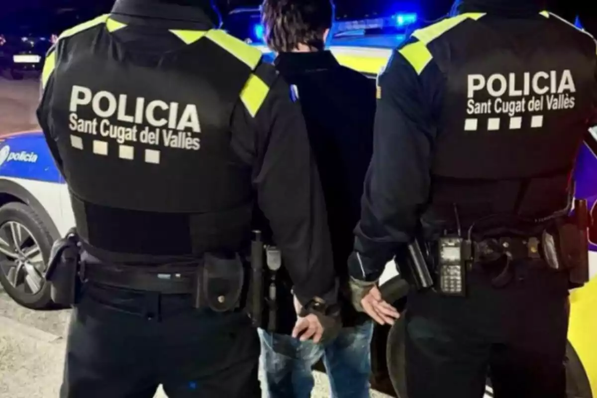 Dos policías de Sant Cugat del Vallès escoltan a una persona esposada frente a un coche patrulla.