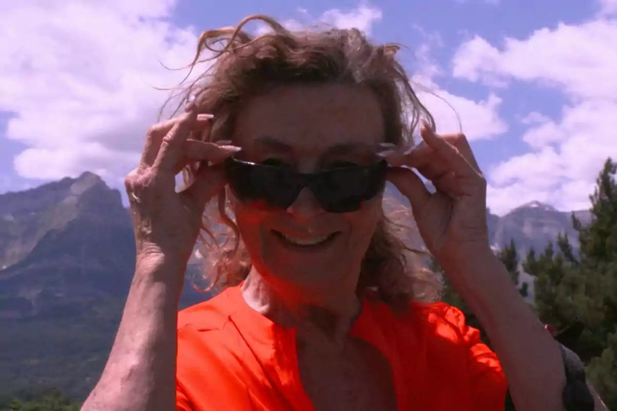 Mujer mayor con gafas de sol y camisa naranja sonriendo al aire libre con montañas y cielo azul de fondo en el programa ¿Por dónde paras? de Aragón TV