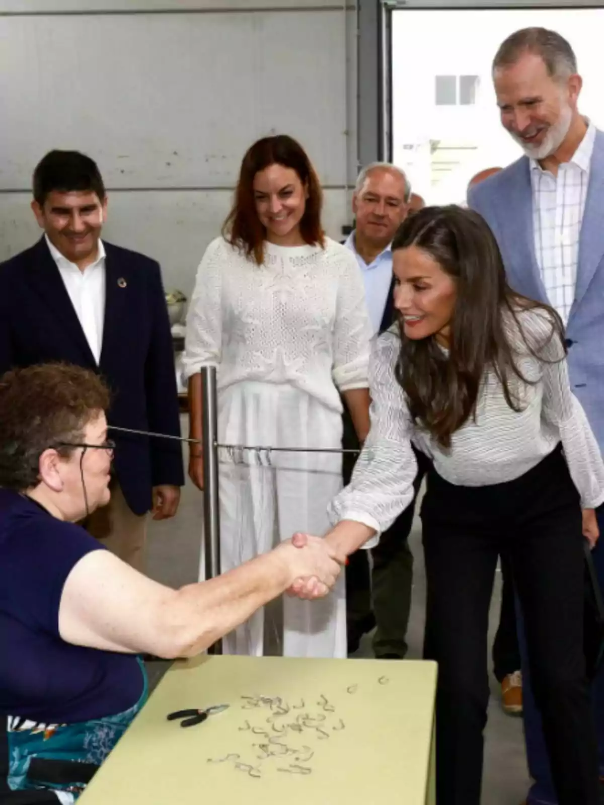 Una mujer sentada en una mesa saluda de mano a la reina Letizia de pie mientras varias personas observan sonrientes en un entorno interior Una mujer sentada en una mesa saluda de mano a la reina Letizia de pie mientras varias personas observan sonrientes en un entorno interior