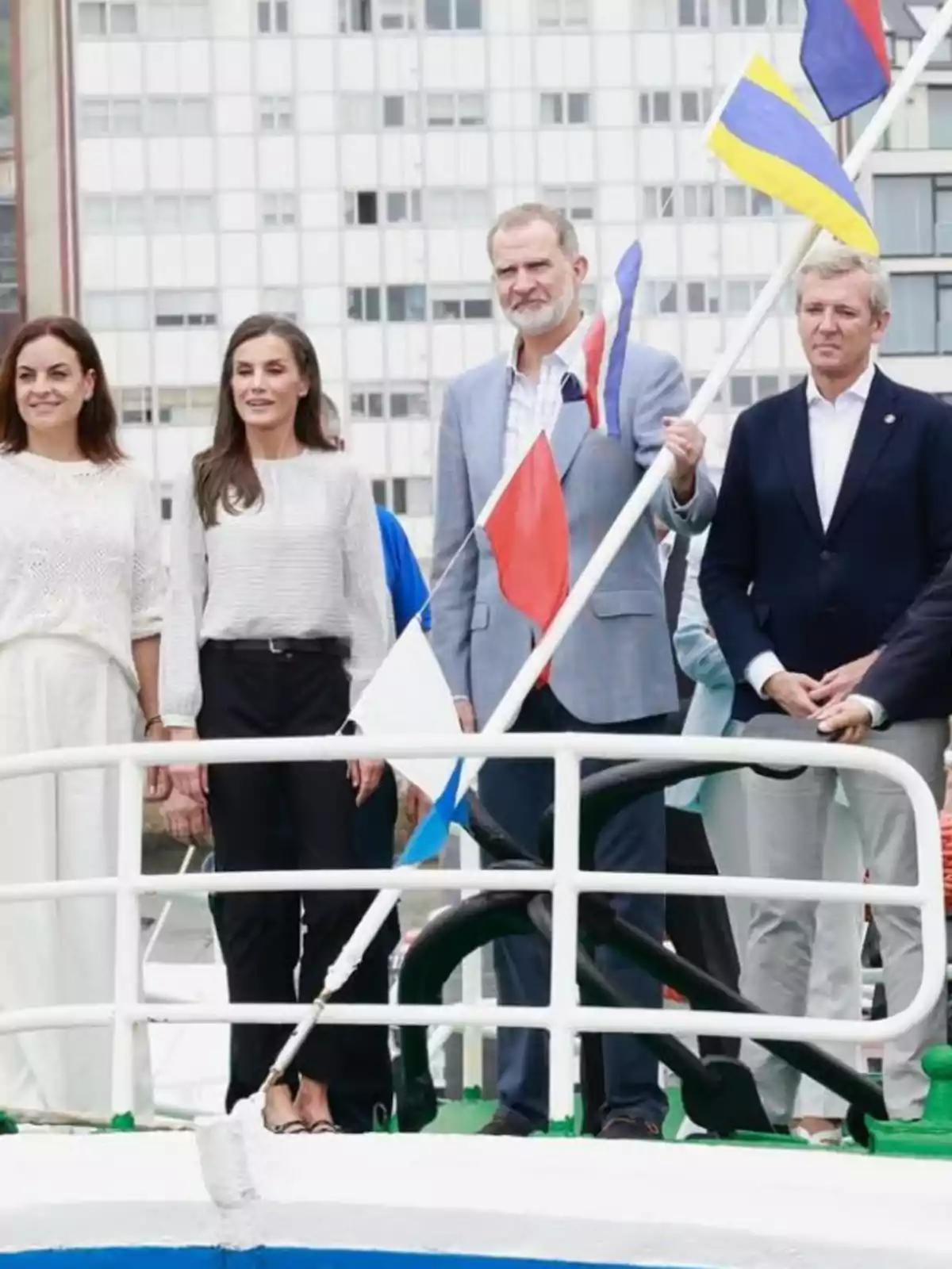 La reina Letizia, Felipe VI y dos personas más posan de pie sobre la cubierta de un barco con varias banderas de colores y un edificio de fondo La reina Letizia, Felipe VI y dos personas más posan de pie sobre la cubierta de un barco con varias banderas de colores y un edificio de fondo