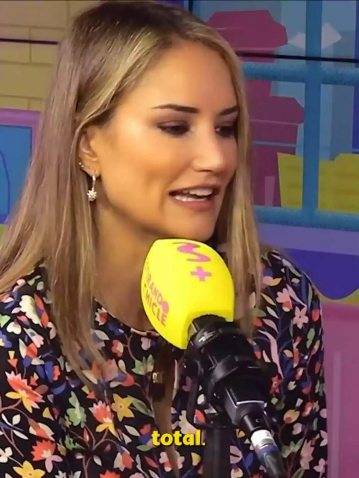 Alba Carrillo con cabello rubio y suelto habla frente a un micrófono amarillo en un estudio colorido, lleva una blusa con estampado floral. Alba Carrillo con cabello rubio y suelto habla frente a un micrófono amarillo en un estudio colorido, lleva una blusa con estampado floral.