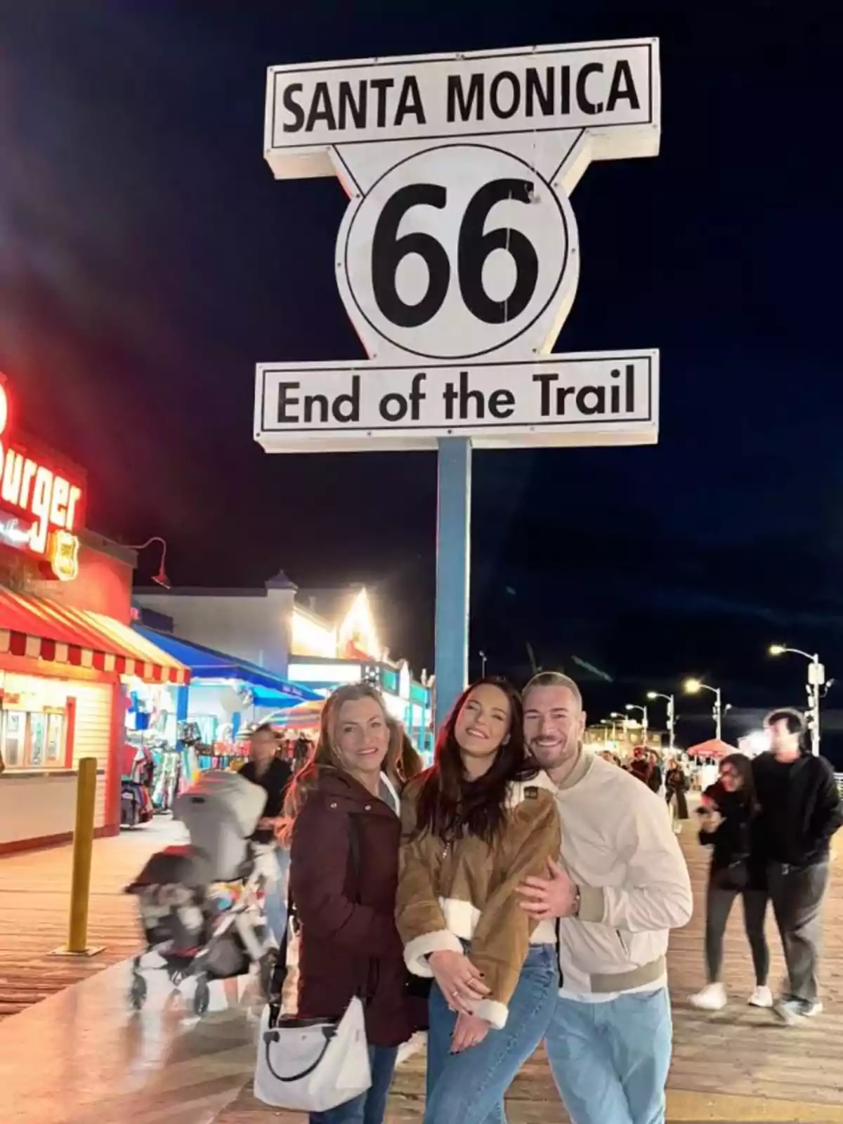 Marta Peñate, Toni Spina y su madre posan sonrientes frente al letrero de la Ruta 66 en Santa Mónica, con luces y tiendas de fondo. Marta Peñate, Toni Spina y su madre posan sonrientes frente al letrero de la Ruta 66 en Santa Mónica, con luces y tiendas de fondo.