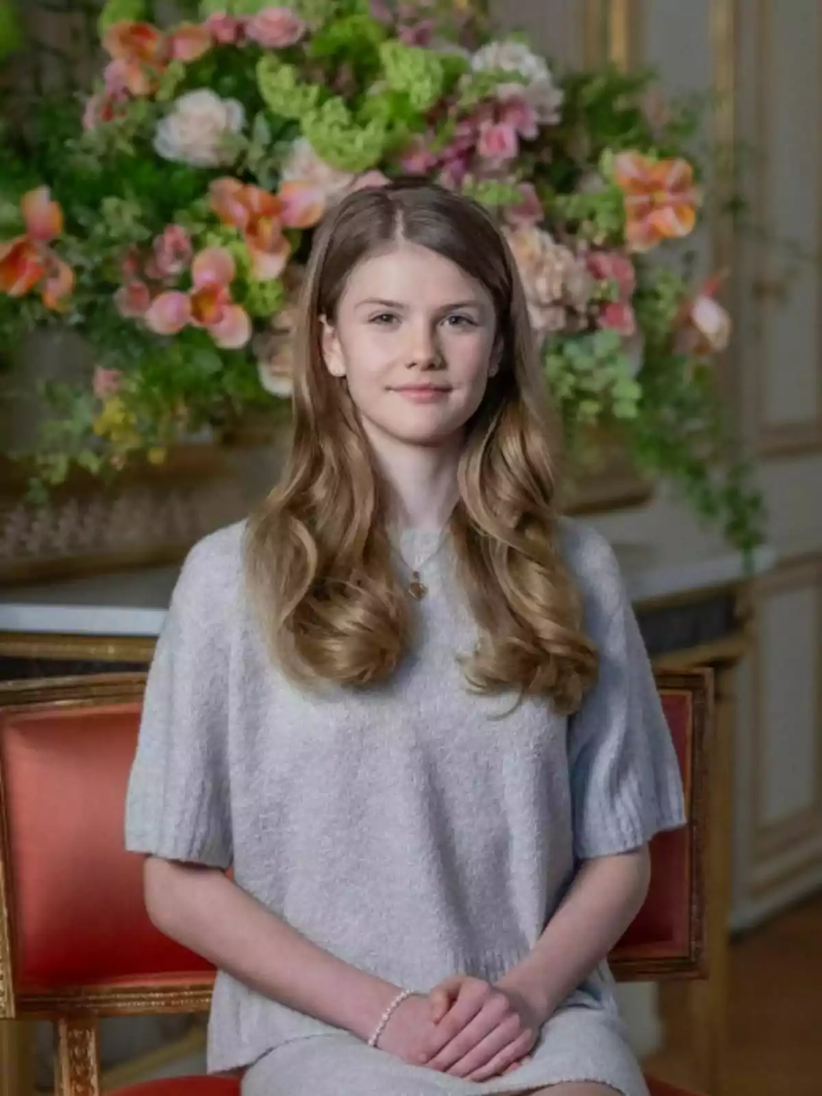La princesa Estelle, vestida con un suéter gris, está sentada en una silla roja frente a un gran arreglo floral colorido. La princesa Estelle, vestida con un suéter gris, está sentada en una silla roja frente a un gran arreglo floral colorido.