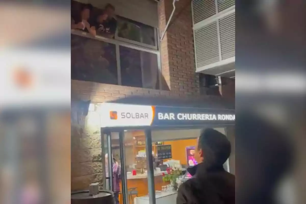 Una persona mira hacia una ventana desde la calle mientras otras personas se asoman desde el interior de un edificio sobre un bar churrería llamado Solbar.