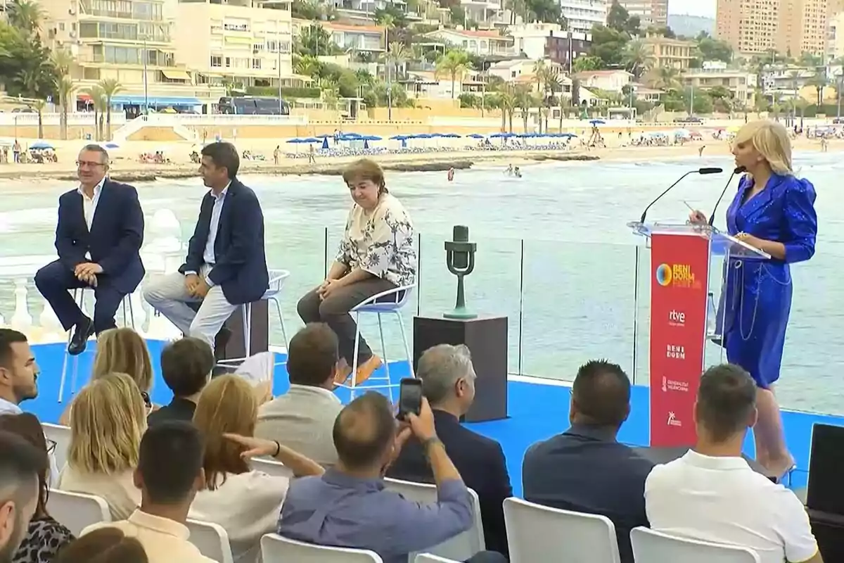 Fotografía de la presentación del Benidorm Fest al aire libre cerca de la playa, con Carlos Mazón, Concepción Cascajosa y Toni Pérez sentados en sillas altas y María Eizaguirre hablando en un podio.