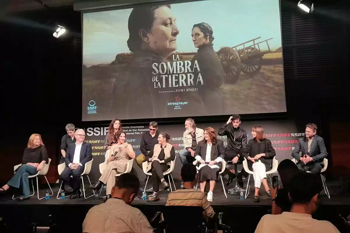 Un grupo de personas como Elvira Mínguez, Adelfa Calvo, María Morales, Ginés García Millán, Montse García o Emilio Sánchez Zaballos está sentado en un escenario en el Festival de San Sebastián durante una conferencia de prensa, con un cartel de la película "La Sombra de la Tierra" proyectado en la pantalla detrás de ellos.