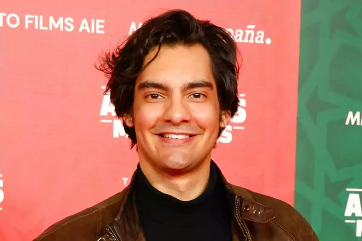 Alejandro Reyes sonriendo con una chaqueta marrón frente a un fondo rojo y verde con texto.