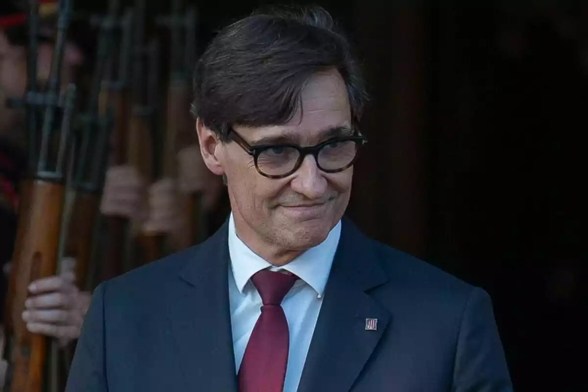 Salvador Illa con traje oscuro, con una corbata roja, sonríe mientras está de pie frente a un grupo de personas que sostienen rifles.