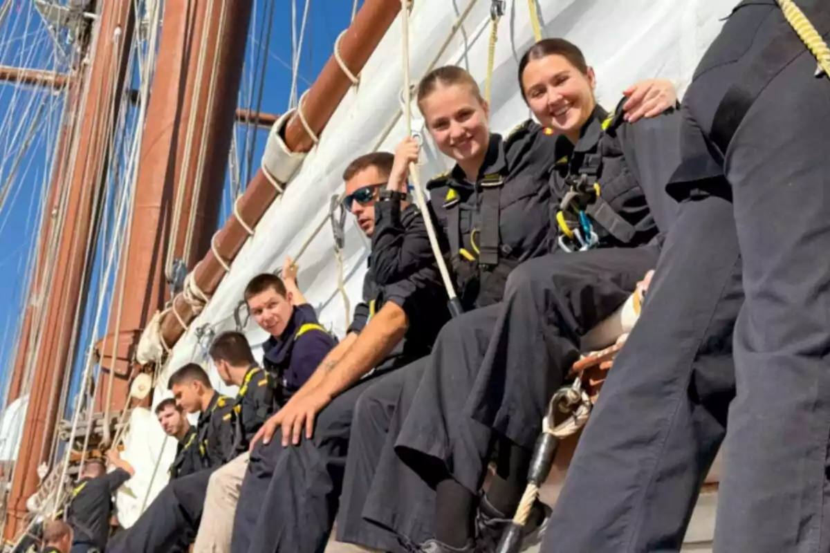 Un grupo de personas vestidas con uniformes oscuros está sentado en la estructura de un barco de vela, sujetándose de cuerdas y sonriendo mientras disfrutan del día soleado. Un grupo de personas vestidas con uniformes oscuros está sentado en la estructura de un barco de vela, sujetándose de cuerdas y sonriendo mientras disfrutan del día soleado.