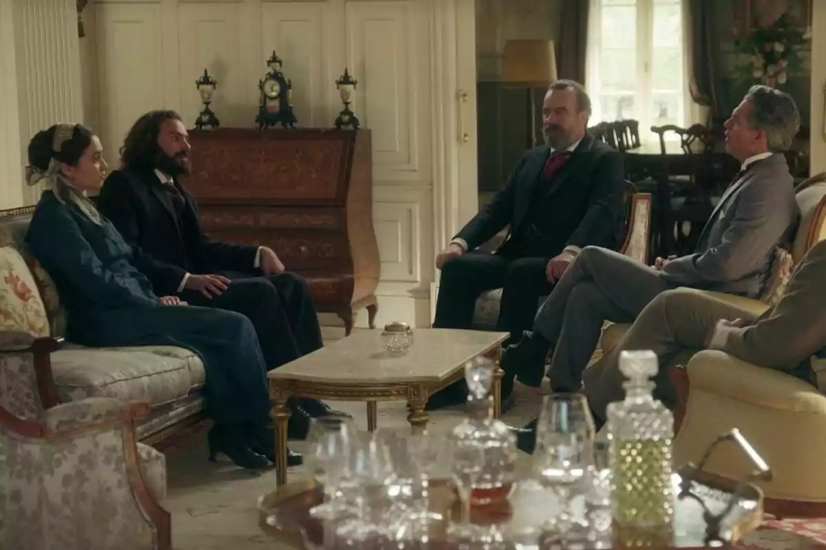 Cuatro personas elegantemente vestidas conversan sentadas en una sala decorada con muebles clásicos y una mesa de centro, al fondo se observa un mueble antiguo y relojes decorativos en La Promesa. Cuatro personas elegantemente vestidas conversan sentadas en una sala decorada con muebles clásicos y una mesa de centro, al fondo se observa un mueble antiguo y relojes decorativos en La Promesa.