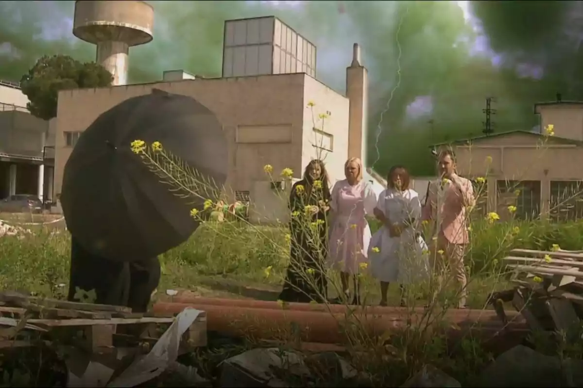Un grupo de personas formado por Inés Hernand, María Patiño, Belén Esteban y Aitor Albizua está de pie en un terreno con vegetación y escombros, mientras una figura con un paraguas negro se encuentra en primer plano; al fondo hay un edificio industrial y el cielo está cubierto de nubes verdes con un rayo visible en una promo de La Familia de la Tele. Un grupo de personas formado por Inés Hernand, María Patiño, Belén Esteban y Aitor Albizua está de pie en un terreno con vegetación y escombros, mientras una figura con un paraguas negro se encuentra en primer plano; al fondo hay un edificio industrial y el cielo está cubierto de nubes verdes con un rayo visible en una promo de La Familia de la Tele.